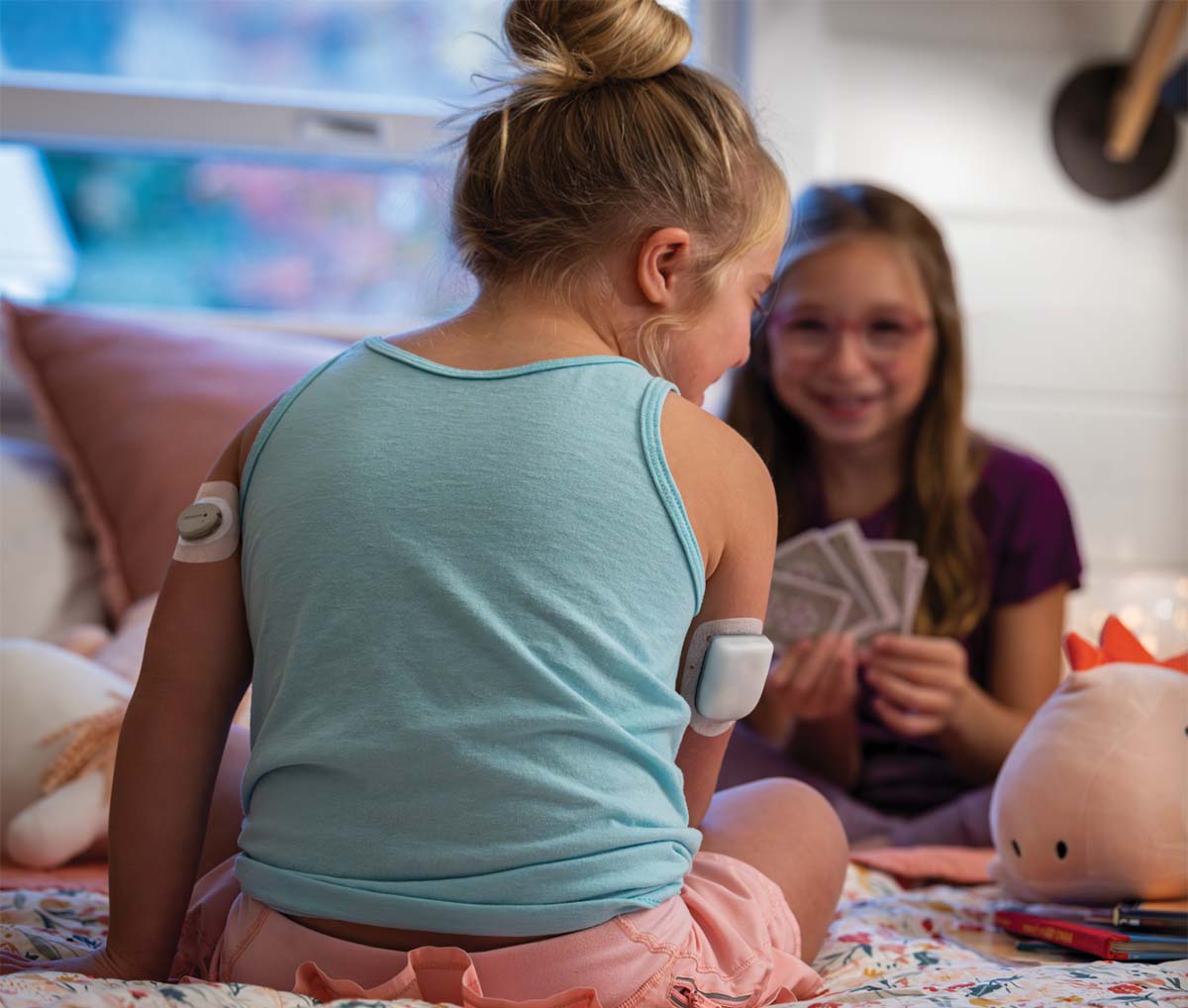 Girls in bedroom playing cards and girl in blue has Omnipod Pod on her right arm and Dexcom G7 on her left arm