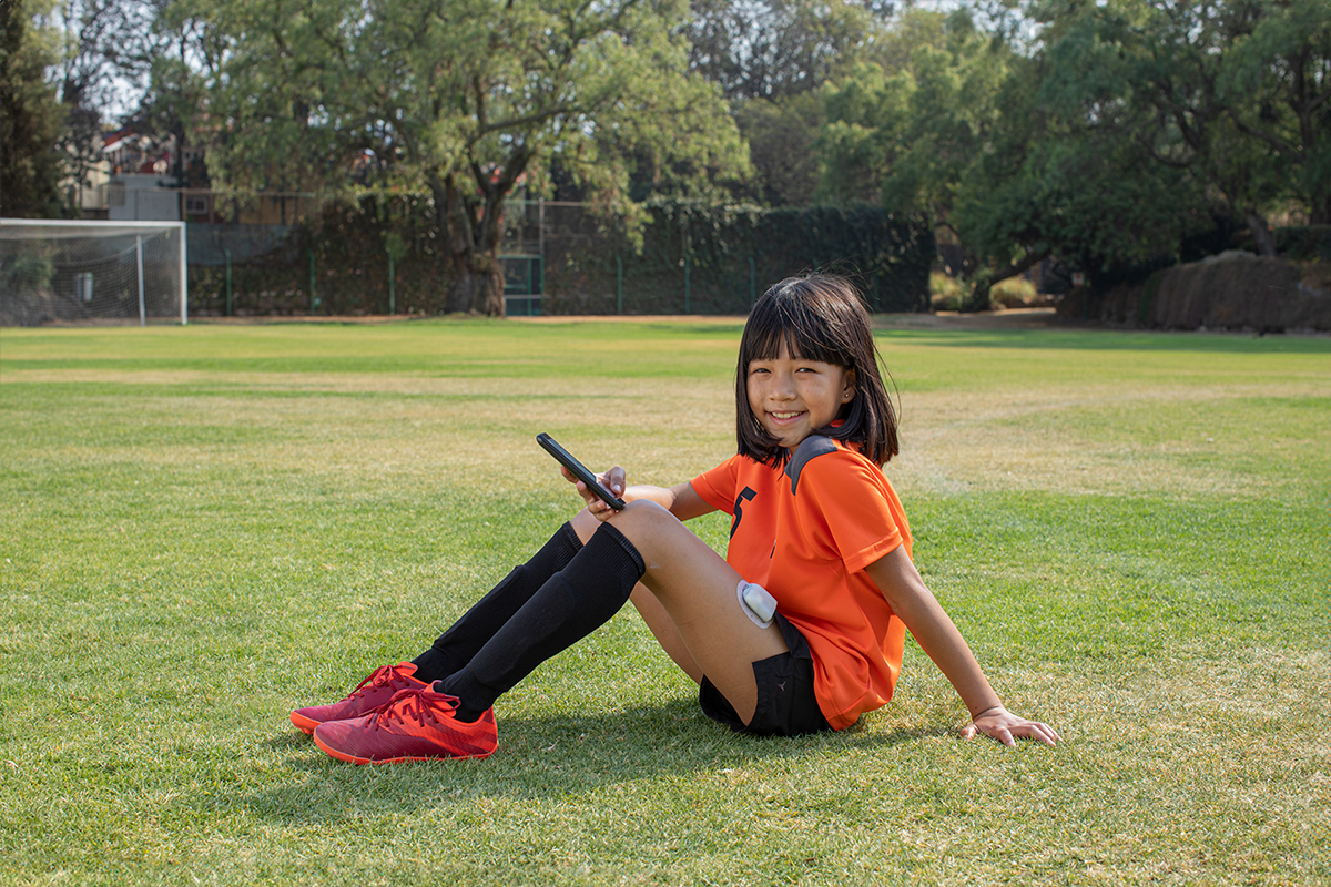 Omnipod Podder young girl in orange soccer uniform sitting down on a soccer field of grass and Pod on her left leg
